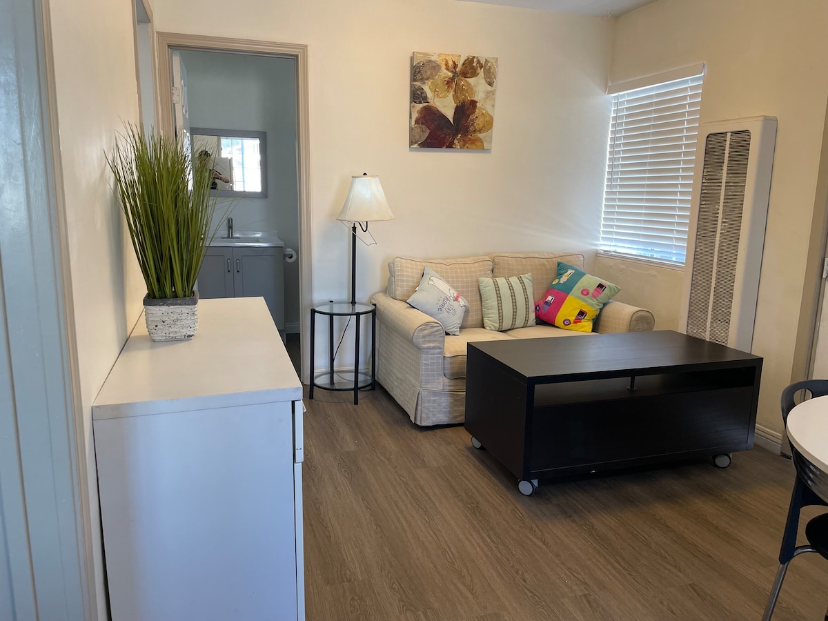 A comfortable living area is depicted, featuring a light-colored sofa adorned with colorful cushions. A coffee table with wheels sits in front of the sofa, and a lamp with a white shade is positioned nearby. Natural light filters through the window, illuminating the space.