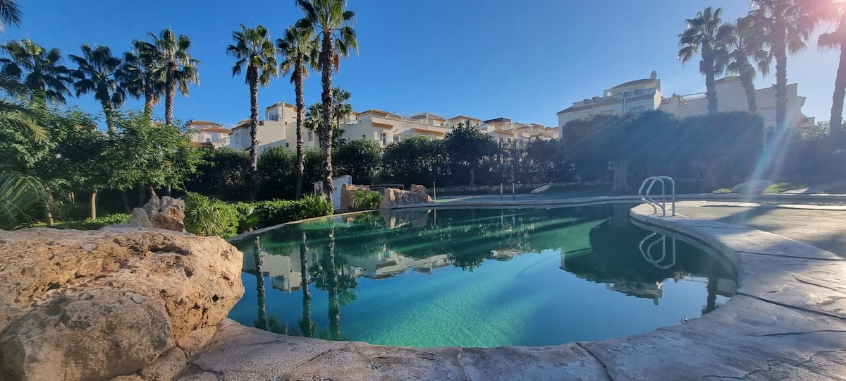 A serene communal pool is framed by lush palm trees and rock features, with clear water reflecting the blue sky. Surrounding villas are visible in the background, providing a peaceful residential ambiance.