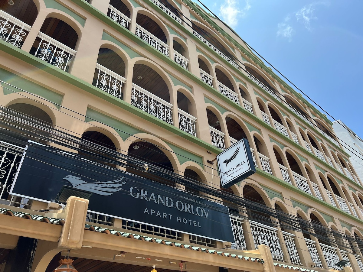 The hotel exterior features a colorful facade with arched windows and intricate ironwork balconies. The signage displays 'Grand Orlov Apart Hotel' prominently. The entrance is shaded by a decorative awning. Multiple stories are visible, highlighting the building's spacious design.