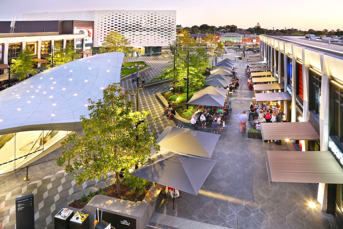 An outdoor shopping area is featured, with modern architecture encompassing a central plaza. Umbrella-covered tables are arranged for seating, while trees provide shade. The space is illuminated with soft lighting, creating an inviting atmosphere for dining and socializing.