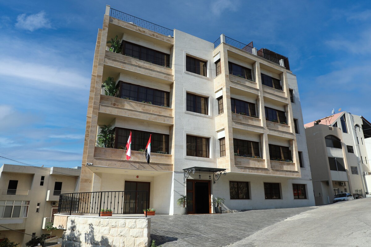The exterior of a modern, multi-story building is showcased against a clear blue sky. Large windows are visible on each floor, framed by neutral-colored walls. A small garden area is located at the entrance, featuring potted plants and flags displayed prominently.