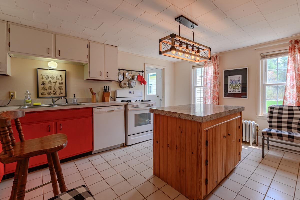 The kitchen showcases red and neutral cabinetry, complemented by a central wooden island. Natural light streams through two windows adorned with sheer curtains. A vintage-style light fixture hangs above the island, while kitchen utensils are neatly organized on the counter. A checkered chair adds a cozy touch.