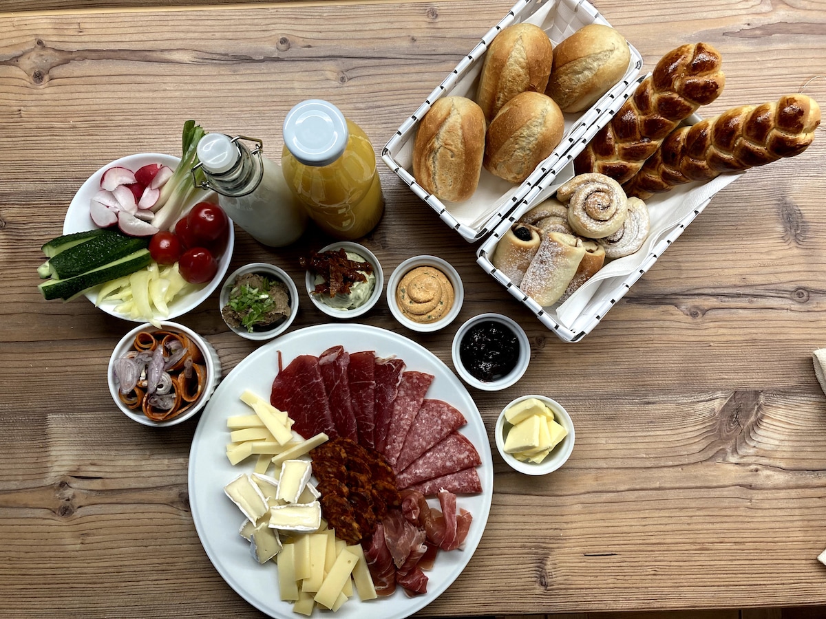 A variety of breakfast items is arranged on a wooden table. A large platter displays an assortment of meats and cheeses, while small bowls hold jams, butter, and pickled vegetables. Fresh bread rolls and pastries are presented in woven baskets alongside bottles of juice and milk.