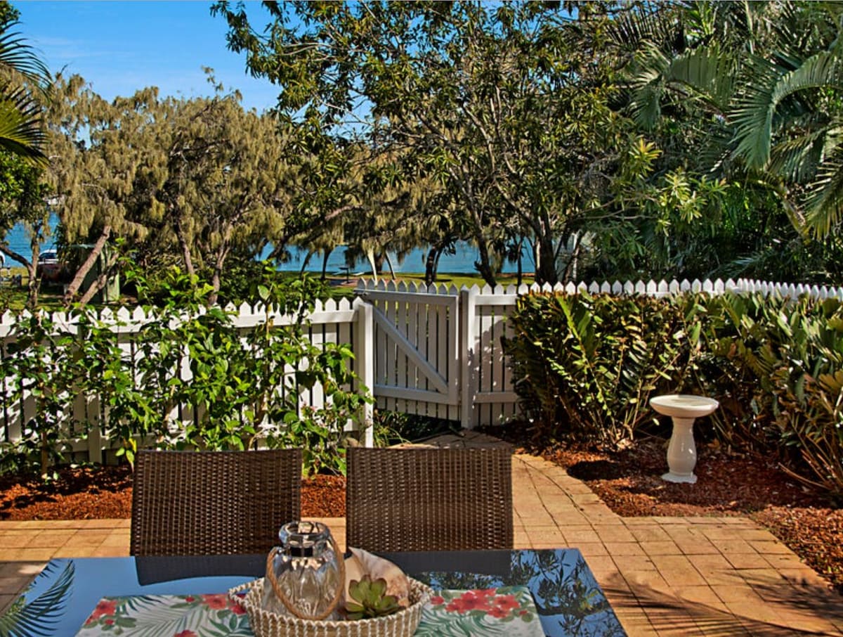 An outdoor patio area features a glass-top dining table surrounded by wicker chairs. Lush greenery lines the space, leading to a white picket fence with a gated entrance. In the background, views of the river are framed by trees under a clear blue sky.