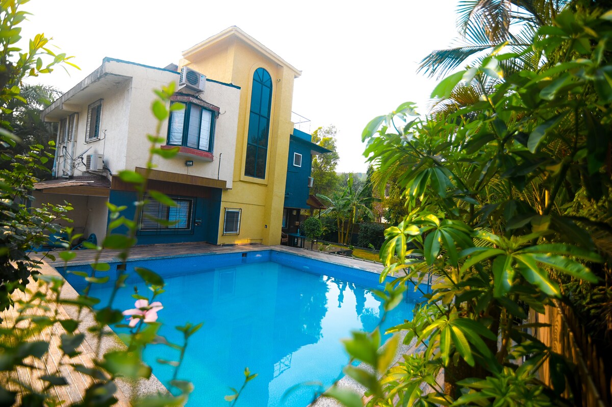 A large swimming pool is surrounded by lush greenery, with the villa prominently featuring an expansive glass window reflecting sunlight. The two-story building displays a charming blend of architectural styles, emphasizing a serene outdoor space equipped for relaxation.