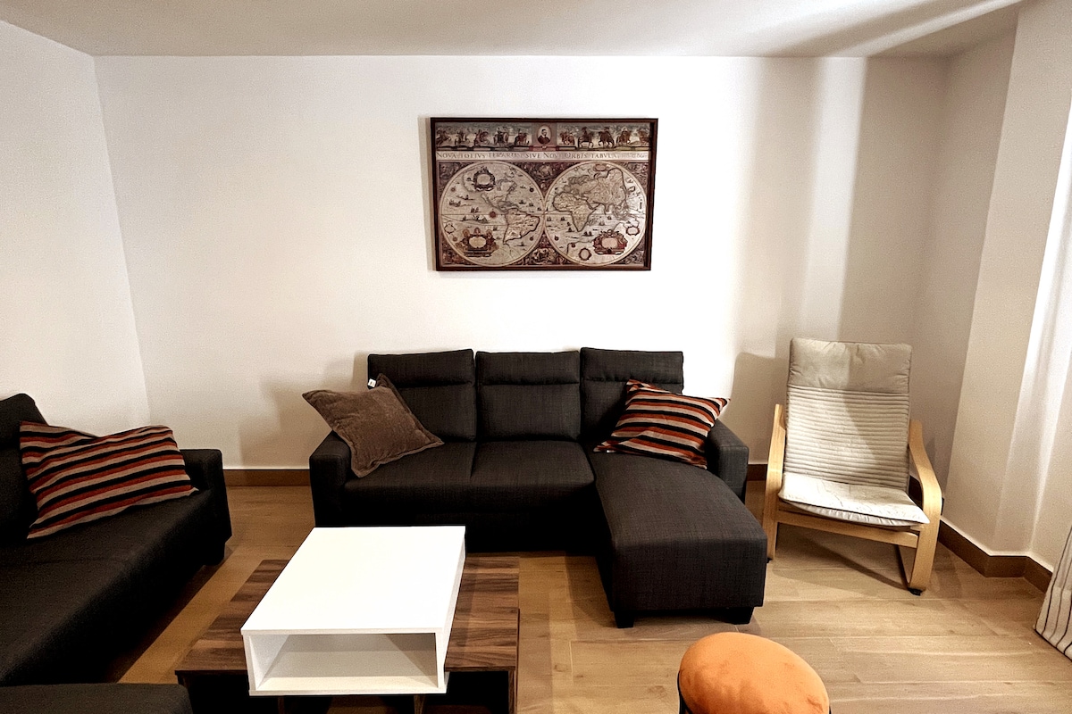 A living area is showcased with a dark, L-shaped sofa adorned with striped cushions. A white coffee table is placed centrally, accompanied by a small orange ottoman. A framed map art piece decorates the wall, complementing the light wood flooring.