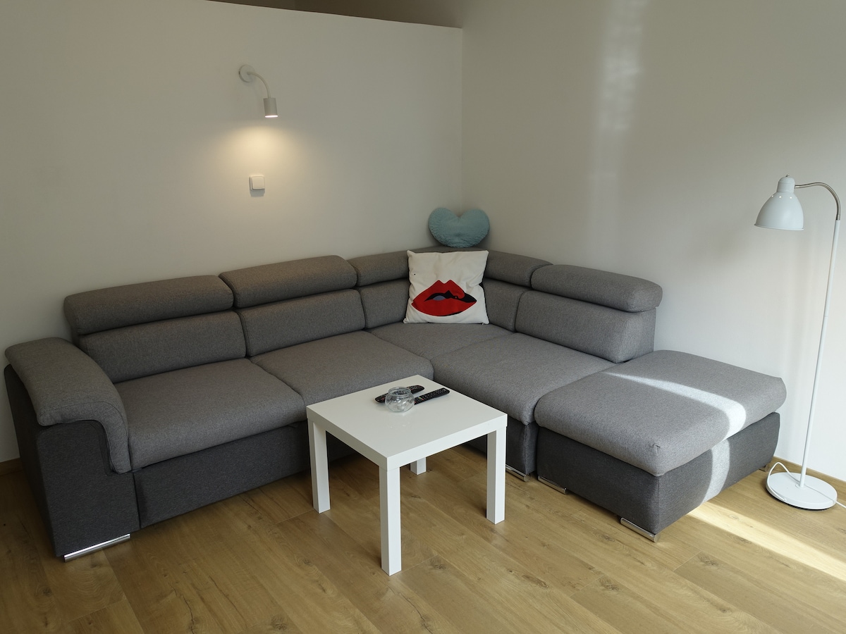 A modern sectional sofa in muted gray is positioned against a white wall, complemented by a matching ottoman. A small white coffee table sits in front, with a remote control and decorative items placed on it. Soft lighting enhances the cozy atmosphere.