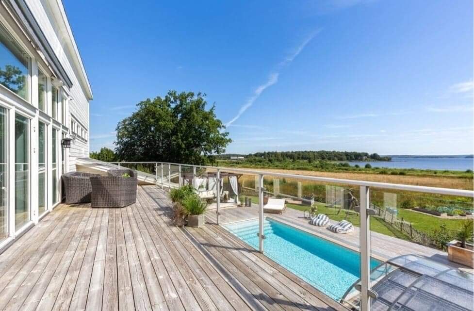 A spacious deck features a clear view of a private pool and is surrounded by lush greenery. Comfortable seating areas are provided with woven furniture. The tranquil landscape includes open fields and a distant waterfront under a bright blue sky.