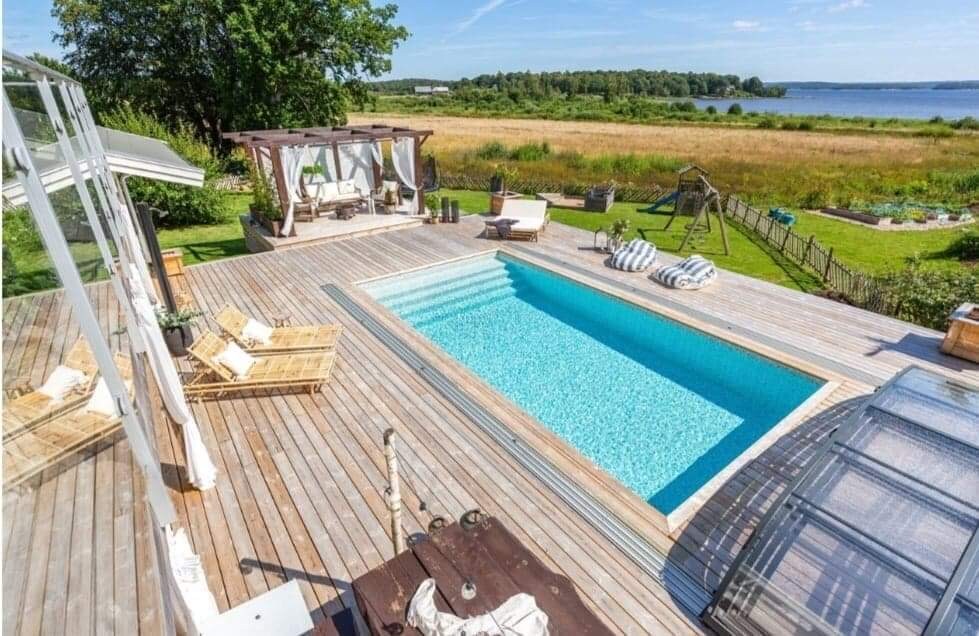 A spacious outdoor area features a swimming pool surrounded by a wooden deck. Comfortable lounge chairs are arranged nearby, and a shaded seating area is visible. Lush greenery and open fields extend towards the water in the background, creating a serene setting.