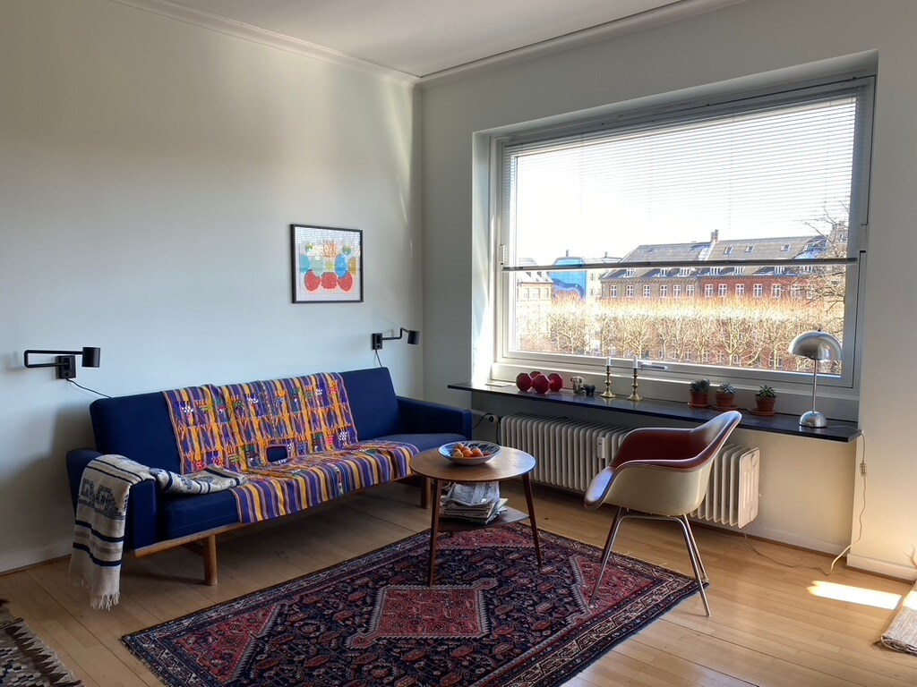 A cozy living area is depicted, featuring a colorful sofa adorned with patterned upholstery. A round coffee table displays a bowl of fruit. Natural light enters through a large window, illuminating the hardwood floor and enhancing the warm atmosphere. A framed art piece adds a touch of personality.