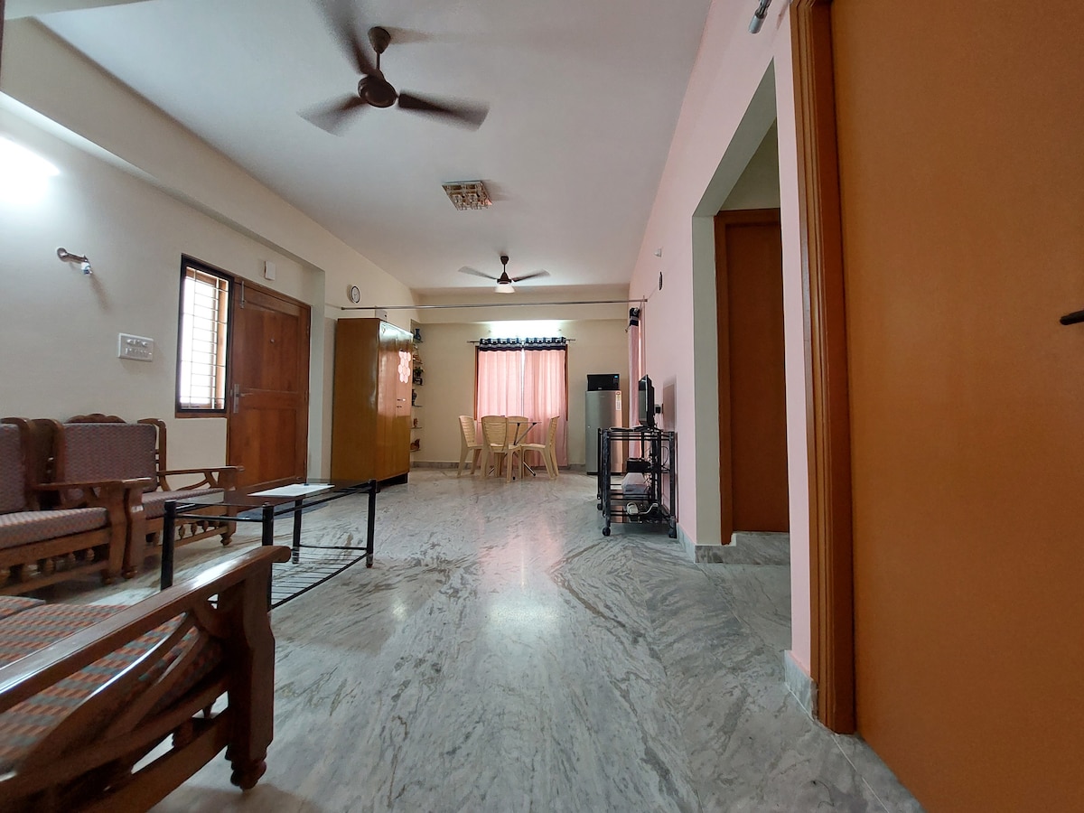 A spacious living area is depicted, featuring a light-colored marble floor. Comfortable seating is arranged near a coffee table, with an adjacent dining area furnished with a round table and chairs. Natural light is filtered through curtains, adding to the inviting ambiance.