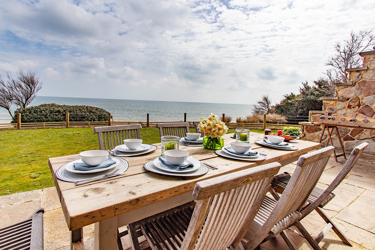 An outdoor dining area features a large wooden table set for a meal, with plates, glasses, and a floral centerpiece. The backdrop includes a view of the sea and lush greenery, with a barbecue nearby and a stone terrace. The sky is partly cloudy, creating a serene atmosphere.