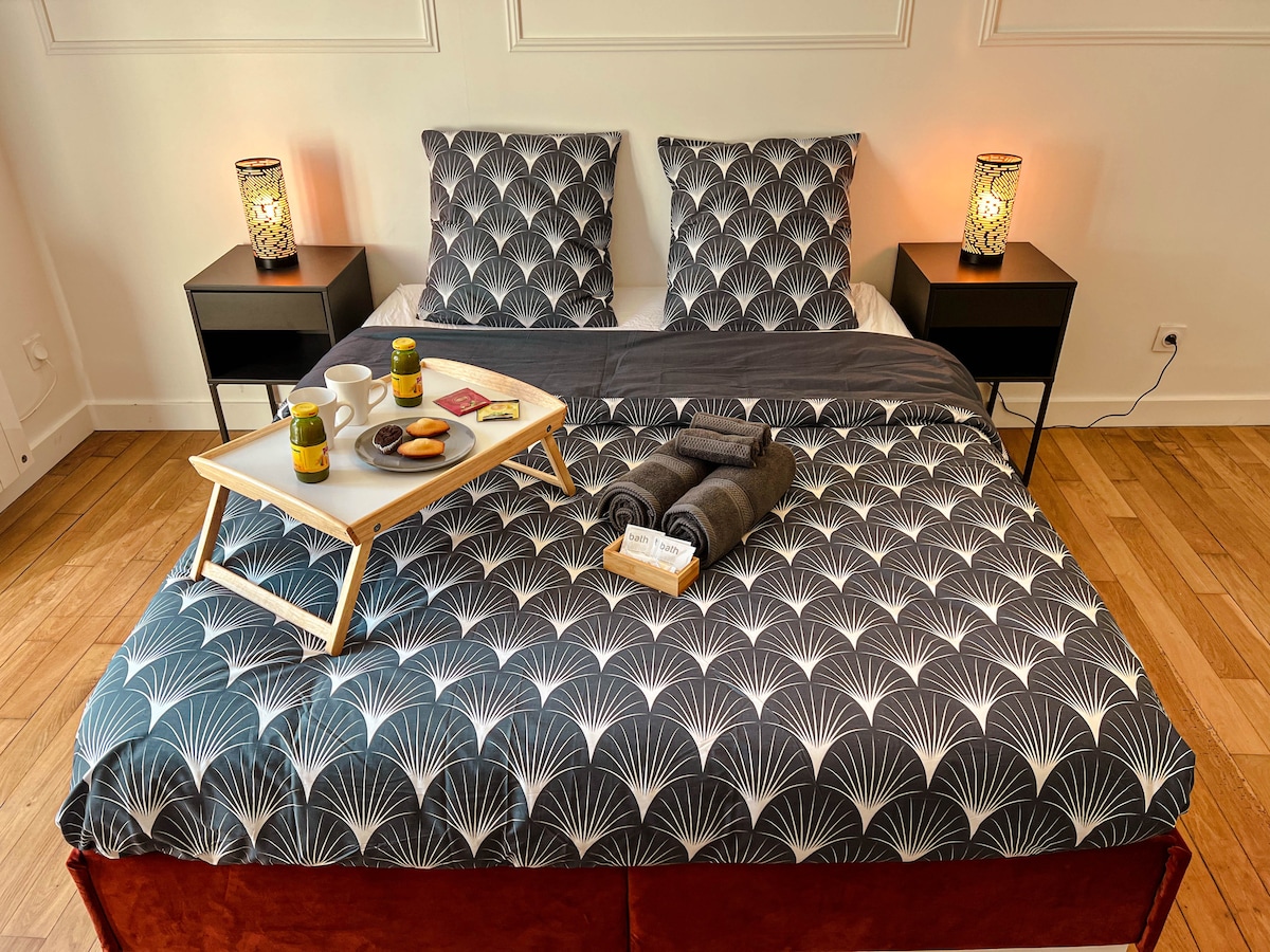 A spacious bed is arranged with patterned bedding and two matching pillows. Nightstands are placed on either side, featuring decorative lamps. A tray with beverages and snacks is set on the bed, alongside neatly rolled towels, enhancing the welcoming ambiance of the room.