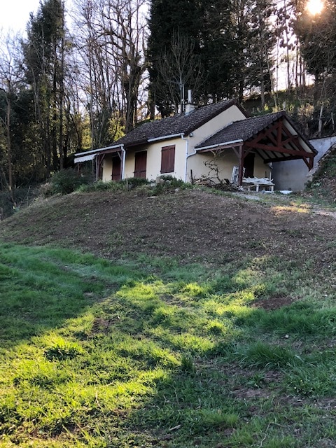 The exterior of the house is presented, featuring a charming facade with wooden shutters and a sloped roof. The property is surrounded by lush greenery and a gentle hill, creating a serene setting. A small outdoor seating area is visible near the entrance.
