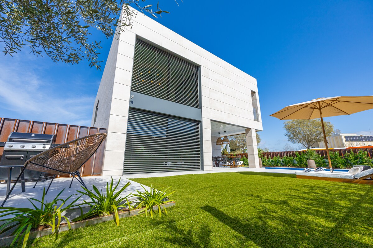 The modern exterior of the villa is framed by a lush green lawn and features a stylish umbrella for shade. A gas grill is visible on the patio, complementing the inviting design of the spacious structure with large windows that bring in natural light.