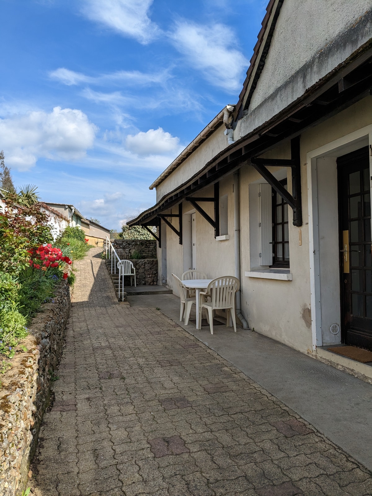 A stone pathway leads to the entrance of the house, flanked by vibrant flowers. Two white chairs and a small table are positioned near the door, inviting outdoor relaxation under the clear blue sky. The exterior of the house features windows and a sleek overhang.