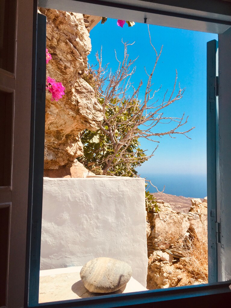 A serene view is framed by open blue shutters, revealing a rocky landscape adorned with sparse vegetation. Bougainvillea flowers add vibrant color, while the expansive blue sky meets the horizon, creating a sense of calm and tranquility.