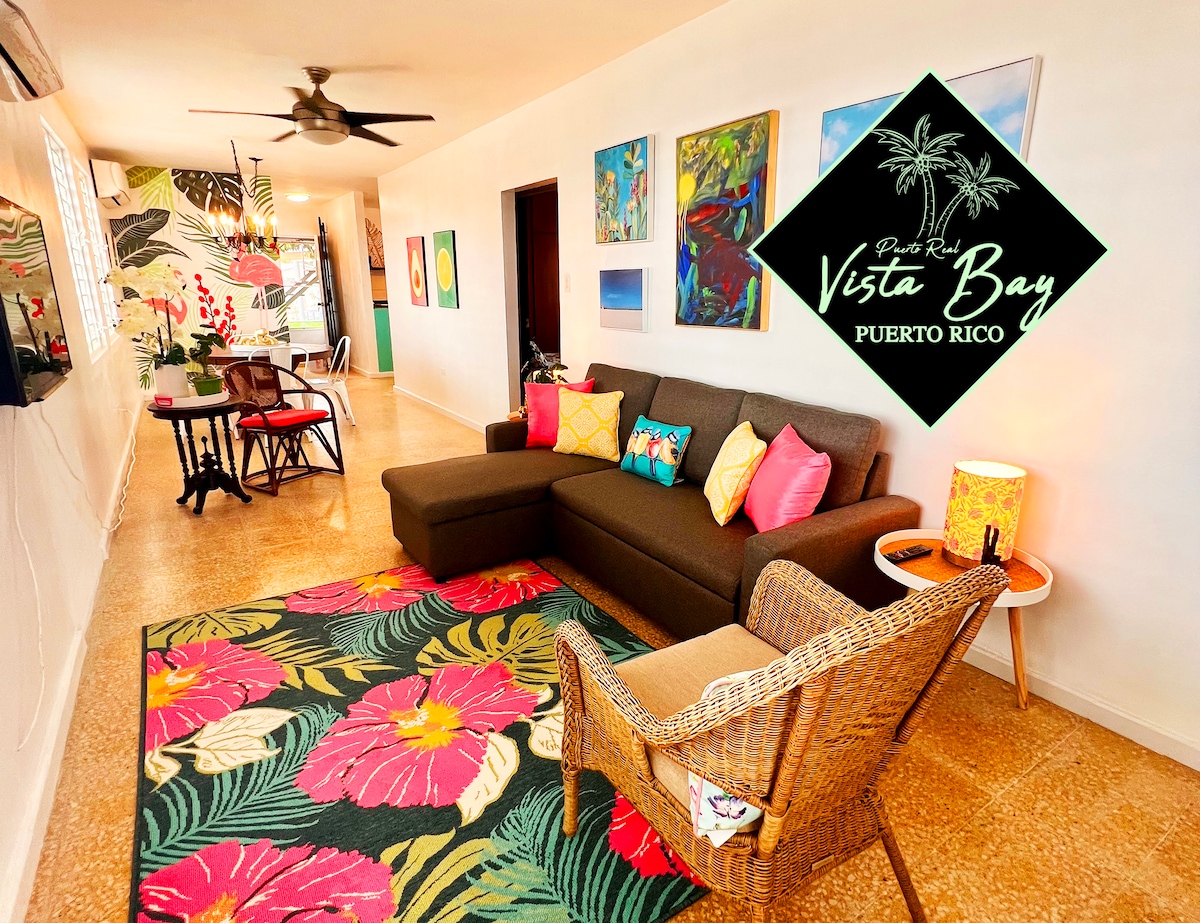 The living area features a comfortable brown sofa with colorful throw pillows and a stylish wicker chair. A vibrant area rug enhances the space, while wall art adds a splash of color. The open layout connects to a dining area with a small table and four chairs.