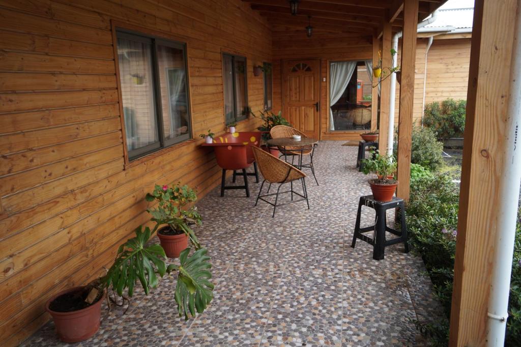A welcoming porch is featured, framed by wooden walls and adorned with several potted plants. Two wicker chairs and a small table create a cozy seating area, while a door leads to the interior space, providing a seamless connection with nature.