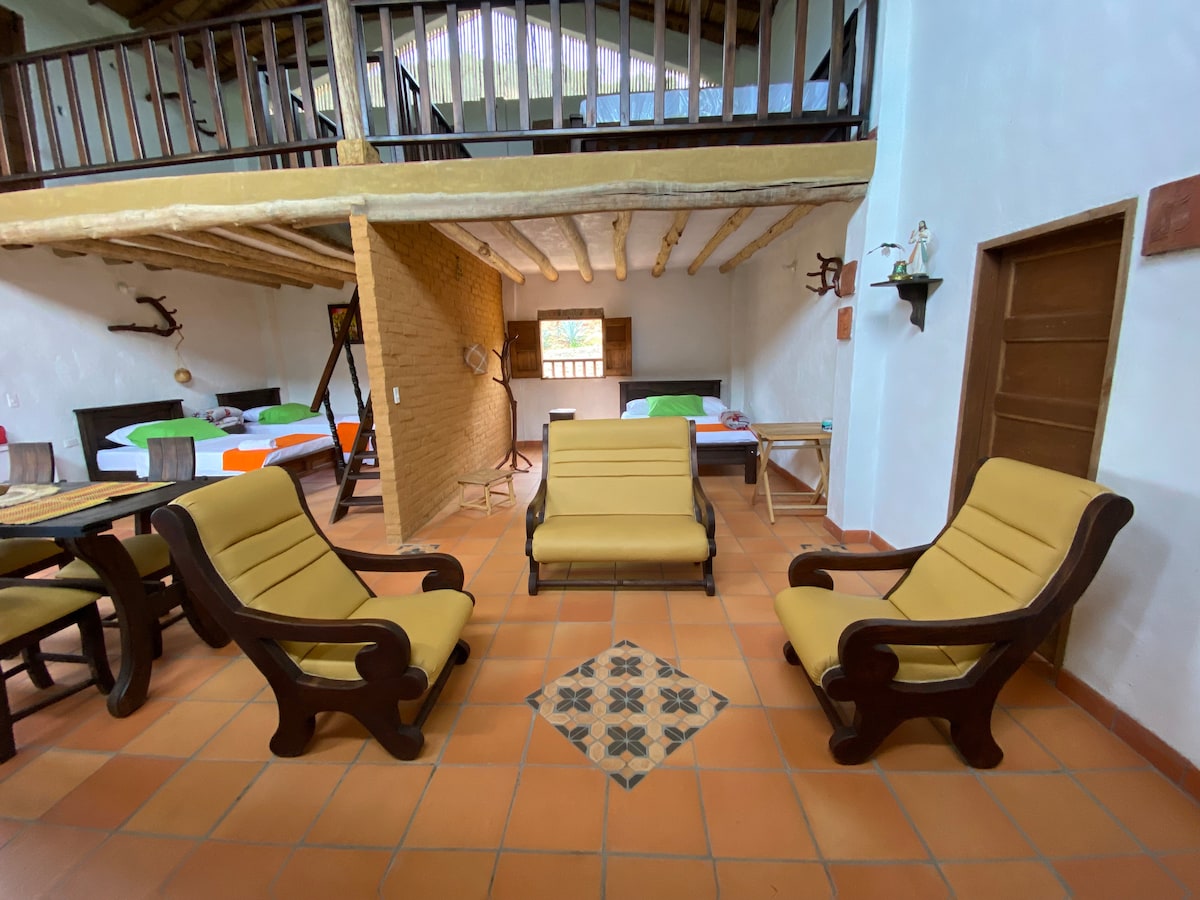A common area features two comfortable lounge chairs in warm tones facing each other. In the background, a lofted sleeping area is visible with neatly made beds and bright bedding. Natural light enters through a window, illuminating the terracotta tiled floor.