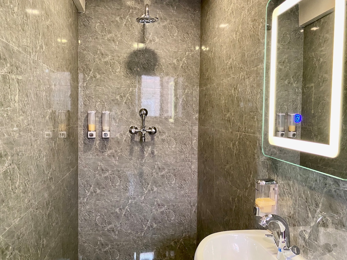 A modern bathroom is presented with sleek gray marble walls and polished fixtures. A stylish mirror with built-in lighting reflects the contemporary design. A washbasin with a round shape is positioned next to a shower fixture, enhancing the clean and organized appearance of the space.