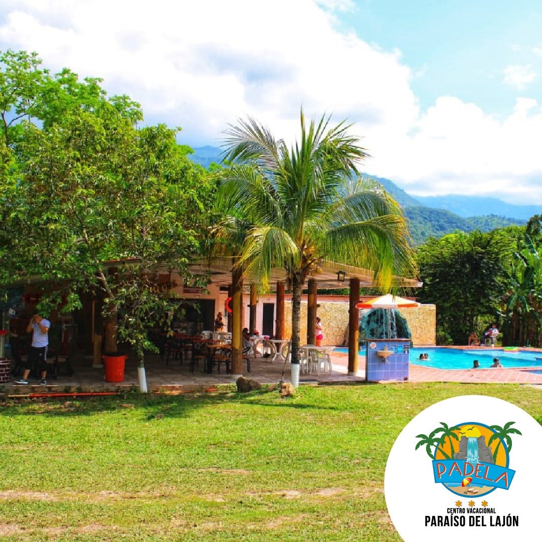 A lush, green lawn is framed by palm trees, leading to a swimming pool area. The inviting space features lounge chairs and a relaxed atmosphere, with a building in the background that accommodates dining and recreational activities. Mountains are visible in the distance under a clear sky.