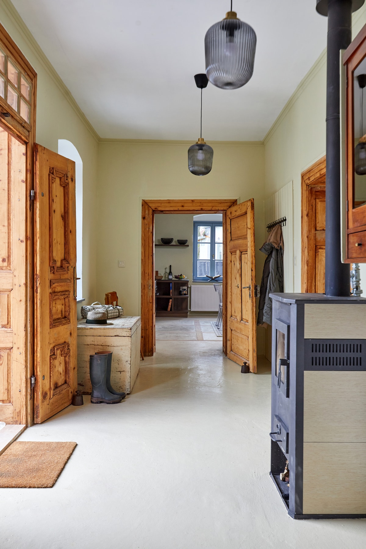 A well-lit hallway features wooden doors and a simple light fixture. A small table is positioned against the wall, accompanied by a few utensils. A wood stove adds warmth to the space. The open entry leads to a cozy kitchen area in the background.