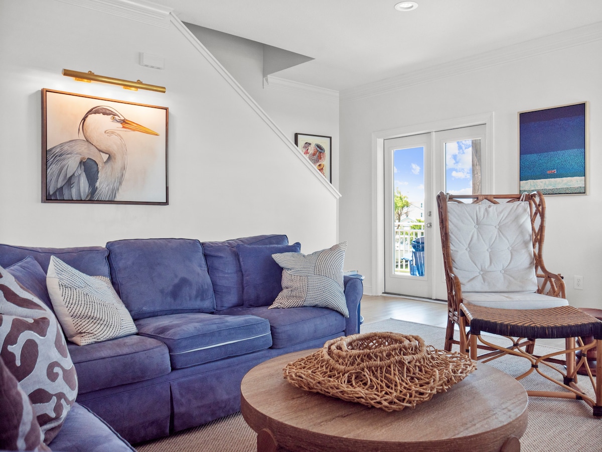 A comfortable living area features a soft blue sectional sofa adorned with patterned throw pillows. A wooden coffee table with a woven centerpiece is positioned in front. Natural light fills the space through glass doors leading to the outdoor deck, enhancing the inviting ambiance.