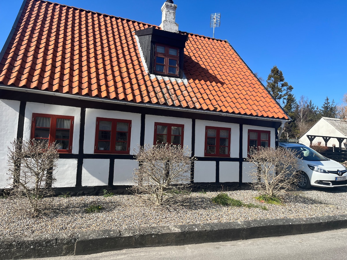 The charming exterior of a two-story house showcases a traditional tiled roof and whitewashed walls with black wooden beams. Prominent red window frames enhance the facade. A gravel yard is visible, featuring understated landscaping, along with a parked vehicle in front.