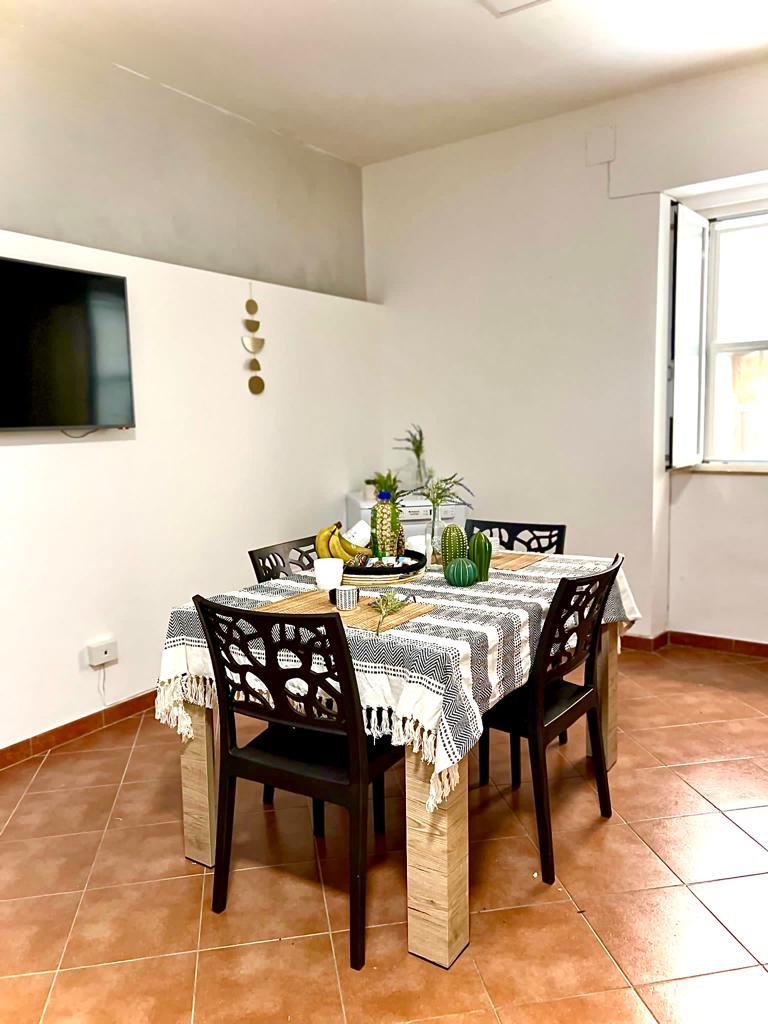 A dining area features a table set with a woven tablecloth, surrounded by four black chairs. A small plant and decorative items are placed on the table. A wall-mounted TV is visible, and natural light enters through an open window.
