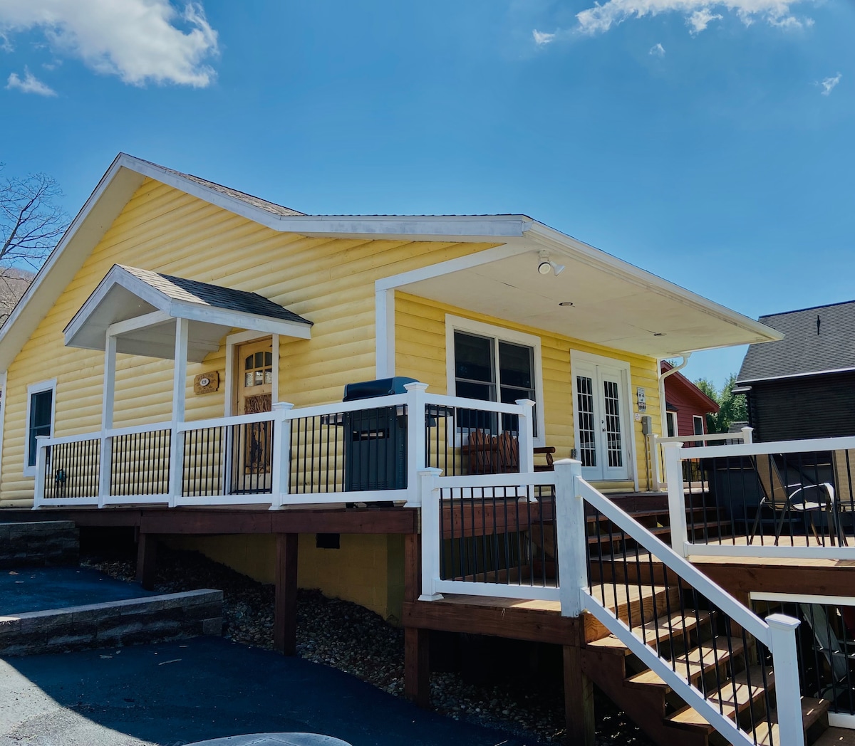 A charming yellow cabin exterior is visible, featuring a spacious deck with seating and steps leading to the entrance. Large windows allow natural light to enter the interior, and a grill is positioned on the deck, indicating a welcoming outdoor space.
