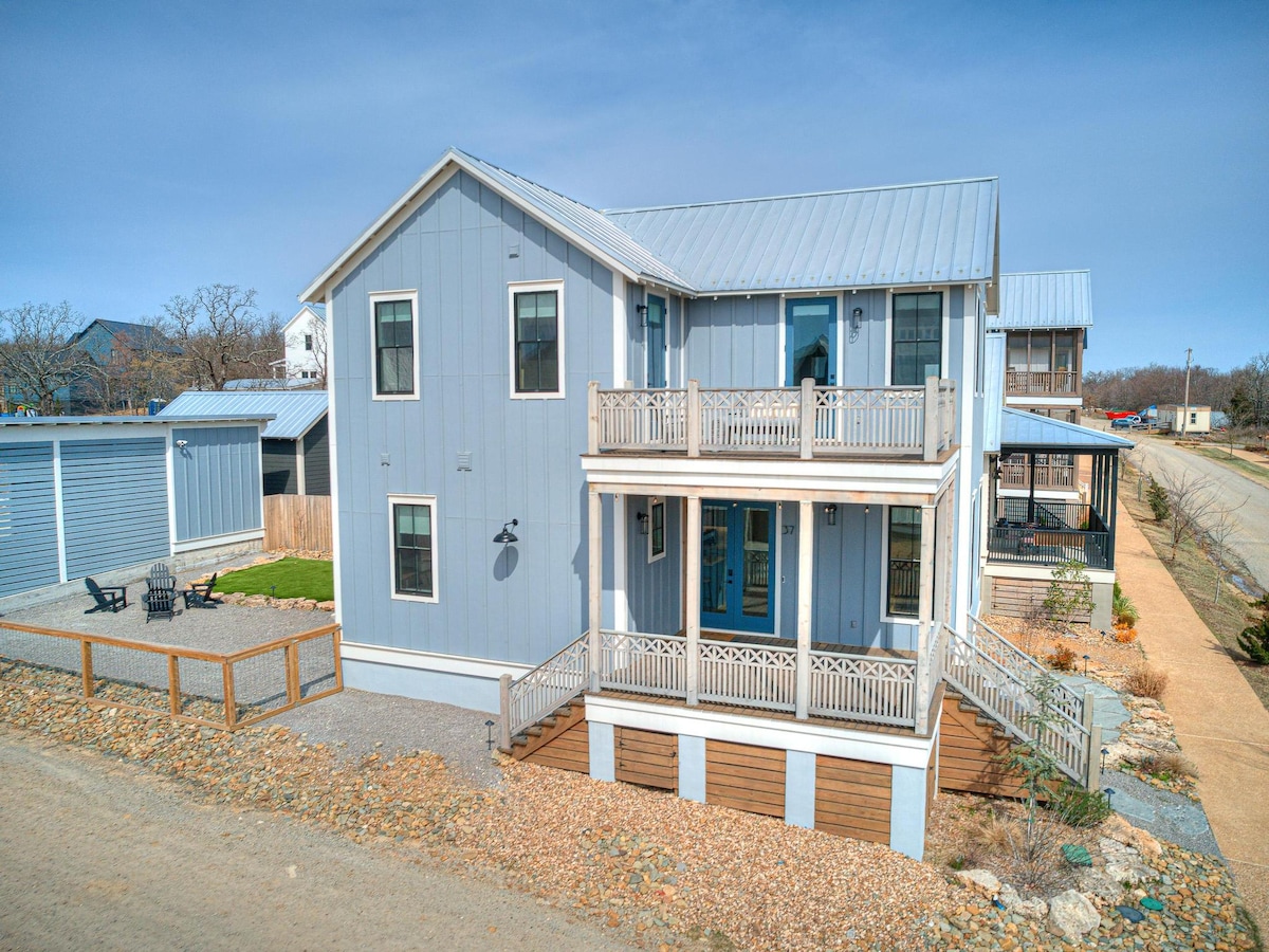 The exterior of the home features a two-story structure with a light blue façade and metal roof. Multiple balconies are visible, with decorative railings, and outdoor seating areas are found at the bottom level. Surrounding landscaping includes gravel pathways and nearby trees.