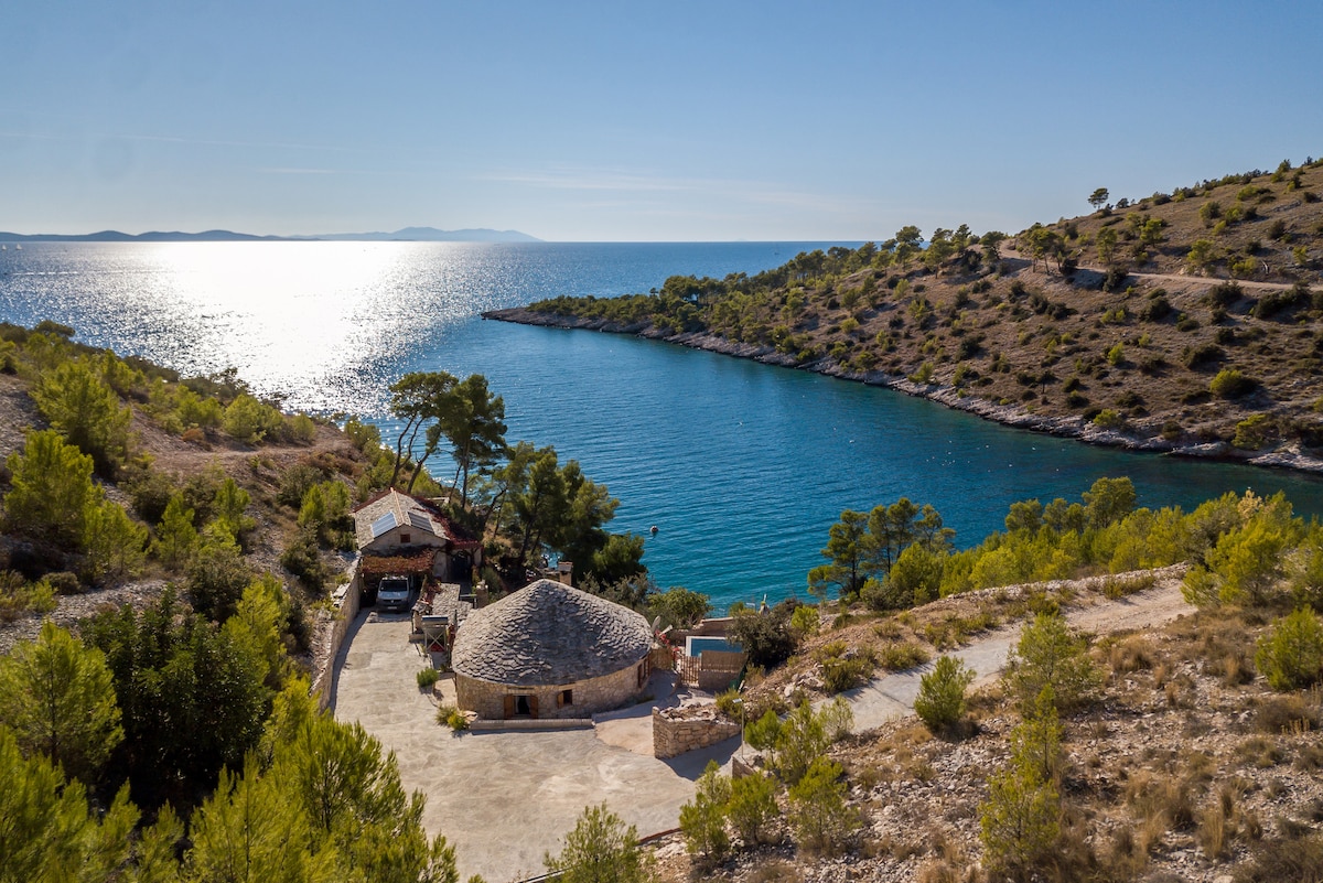 Aerial view captures The Old Captain's House nestled in a serene cove, surrounded by natural greenery. The bright, sparkling sea reflects sunlight, while the rustic structure features a distinctive round roof. The tranquil landscape offers a sense of seclusion and connection to nature.
