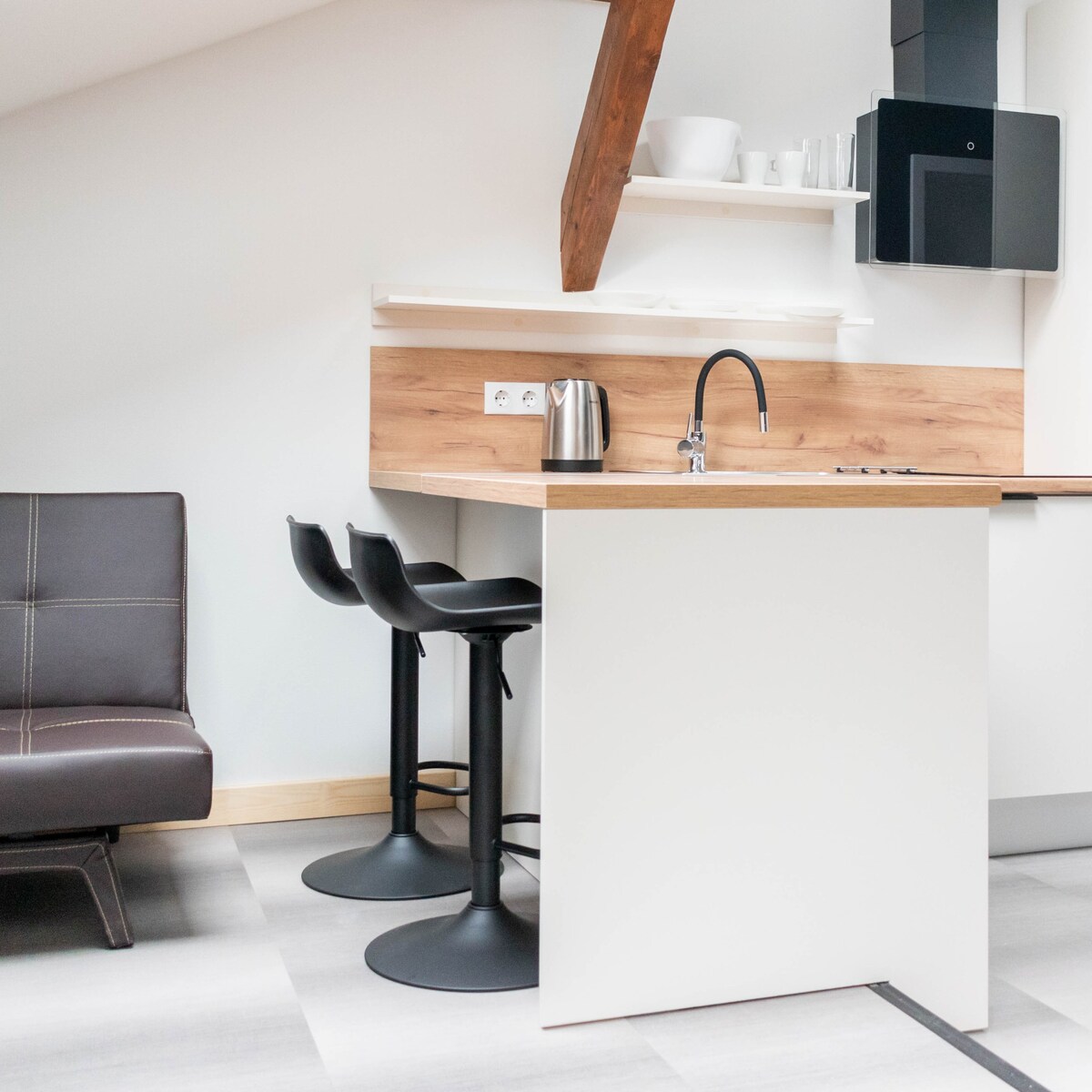 A compact kitchen area is combined with a modern dining space, featuring sleek black bar stools at a light wood counter. The counter is equipped with a kettle and stove, framed by wooden beams that add architectural interest to the space.