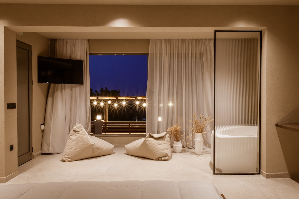 A serene interior space features a king-size bed with soft linens, located near a large window with sheer curtains allowing light to filter through. Cozy bean bags are placed in the corner, and a glass enclosure reveals a stylish indoor Jacuzzi beside a balcony view.