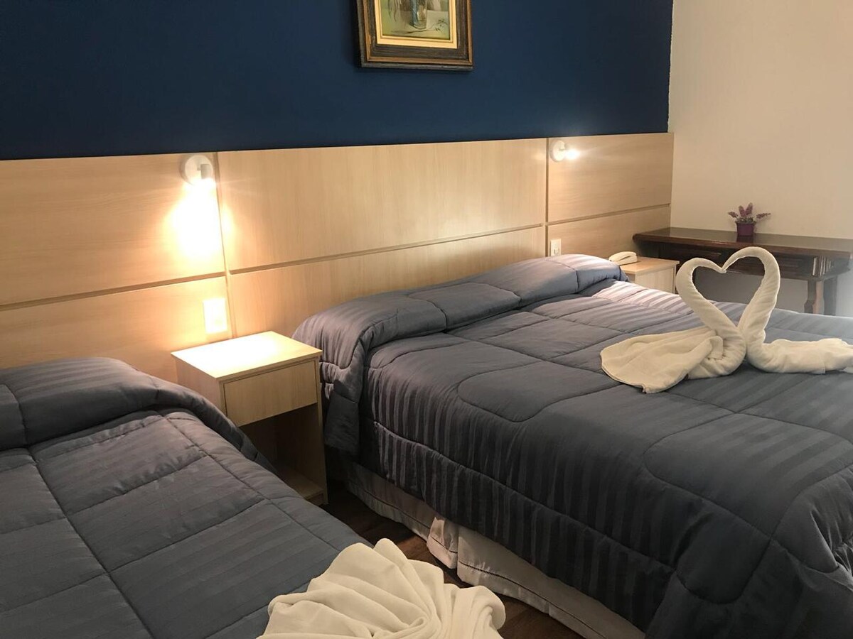 A room is highlighted by two spacious beds, each with a soft, gray comforter. Elegant towel art in the shape of a heart is arranged on one bed. A nightstand with a lamp and a painting above adds a touch of simplicity and warmth to the space.