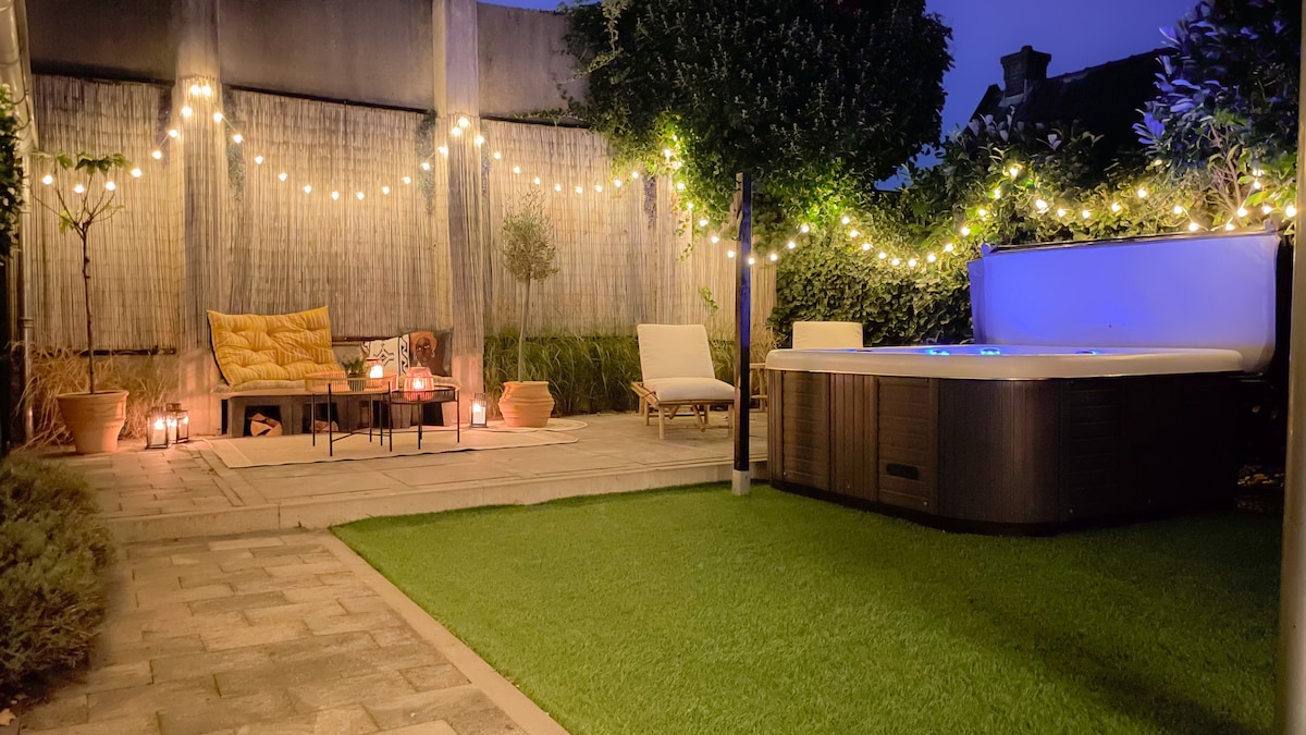 A secluded outdoor space is presented, featuring a jacuzzi surrounded by soft lighting and greenery. Comfortable seating arrangements, including a lounge chair and a sofa, are visible. Decorative plants and cozy elements create a warm ambiance in this private garden setting.
