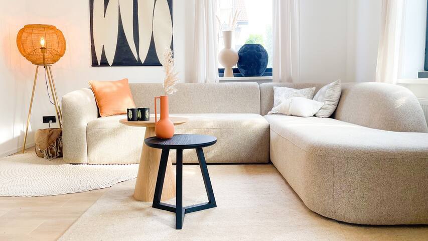 A light-filled living area is presented with a curved, neutral-toned sofa and a round black side table. A textured rug covers the wooden floor, while a floor lamp adds warmth. Decorative elements include a vase and artwork on the wall, complementing the modern aesthetic.