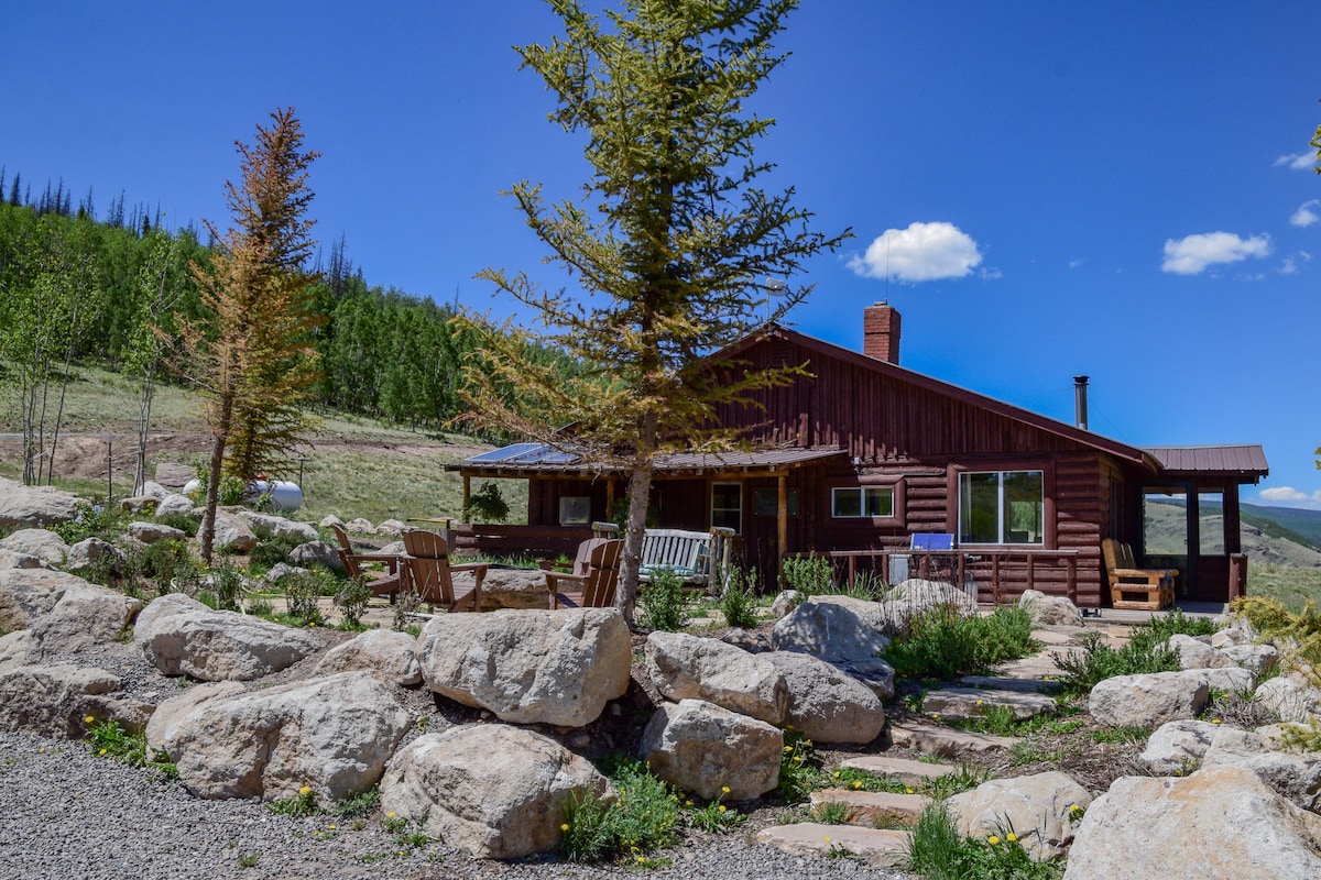 A rustic cabin is nestled among large rock formations, with a few trees providing natural landscaping. Wooden chairs and a table are positioned on a gravel area, inviting outdoor relaxation. The structure features large windows that offer expansive views of the surrounding mountains and sky.