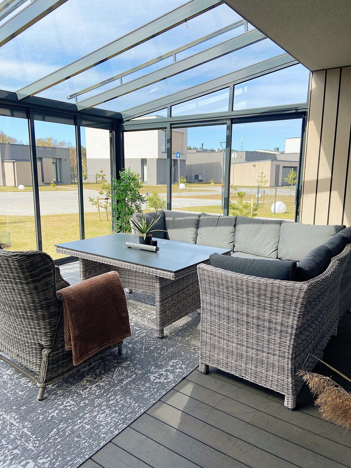 A sunlit terrace area is enclosed with glass, featuring a comfortable seating arrangement with gray cushions and a coffee table. Green plants are placed around the space, offering a relaxing environment with a view of the surrounding area.