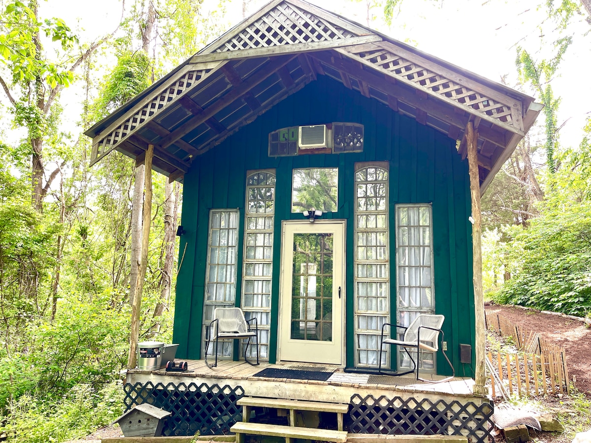 A charming cabin features a dark green exterior, highlighted by large, floor-to-ceiling windows that invite natural light. The covered porch is furnished with two chairs, offering a cozy spot to enjoy the surrounding greenery and tranquility of the wooded setting.