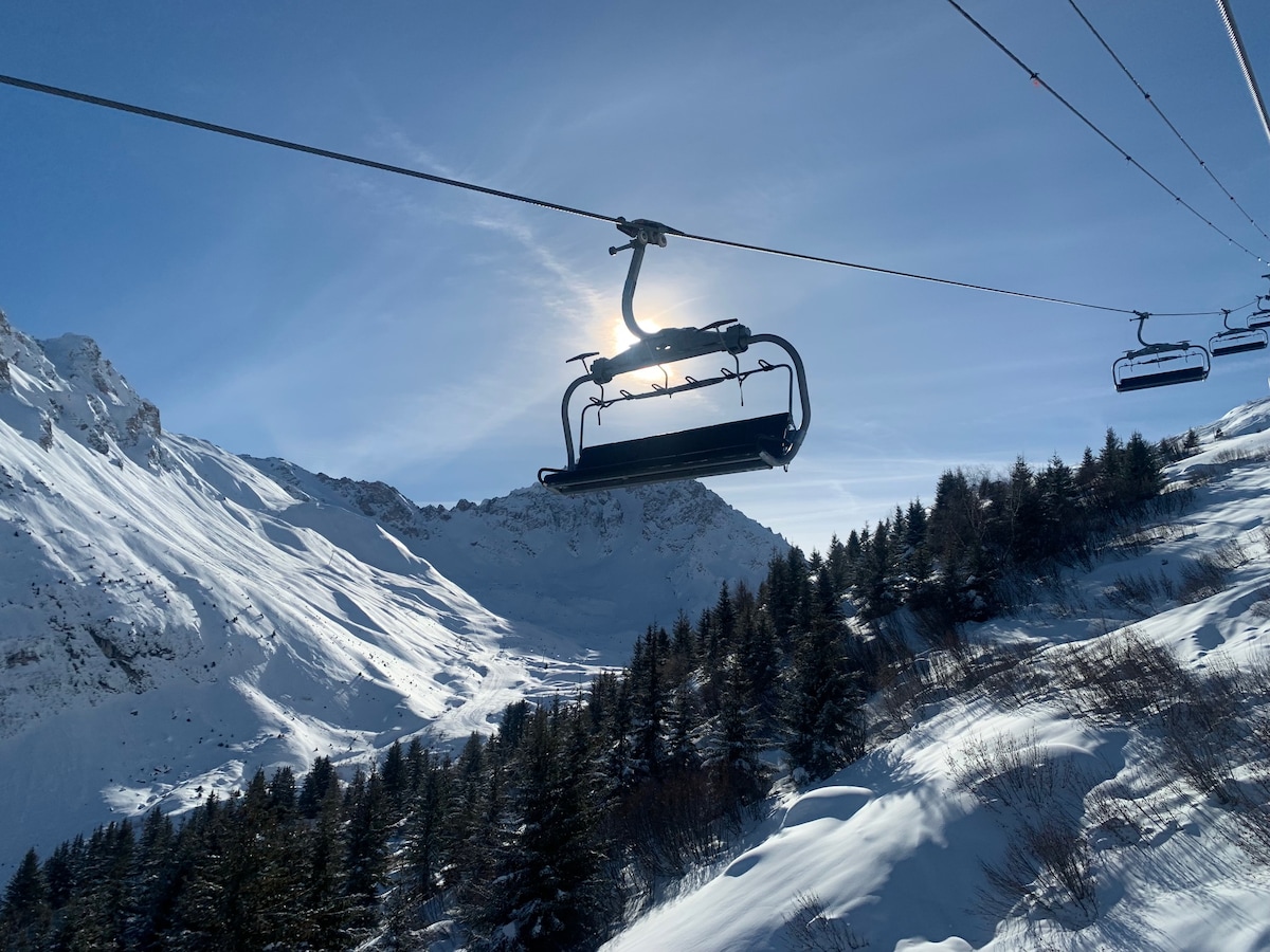 A ski lift is seen ascending against a clear blue sky, with sunlight creating a halo effect around its silhouette. Snow-covered mountains rise in the background, while evergreen trees dot the foreground, indicating a picturesque winter landscape.