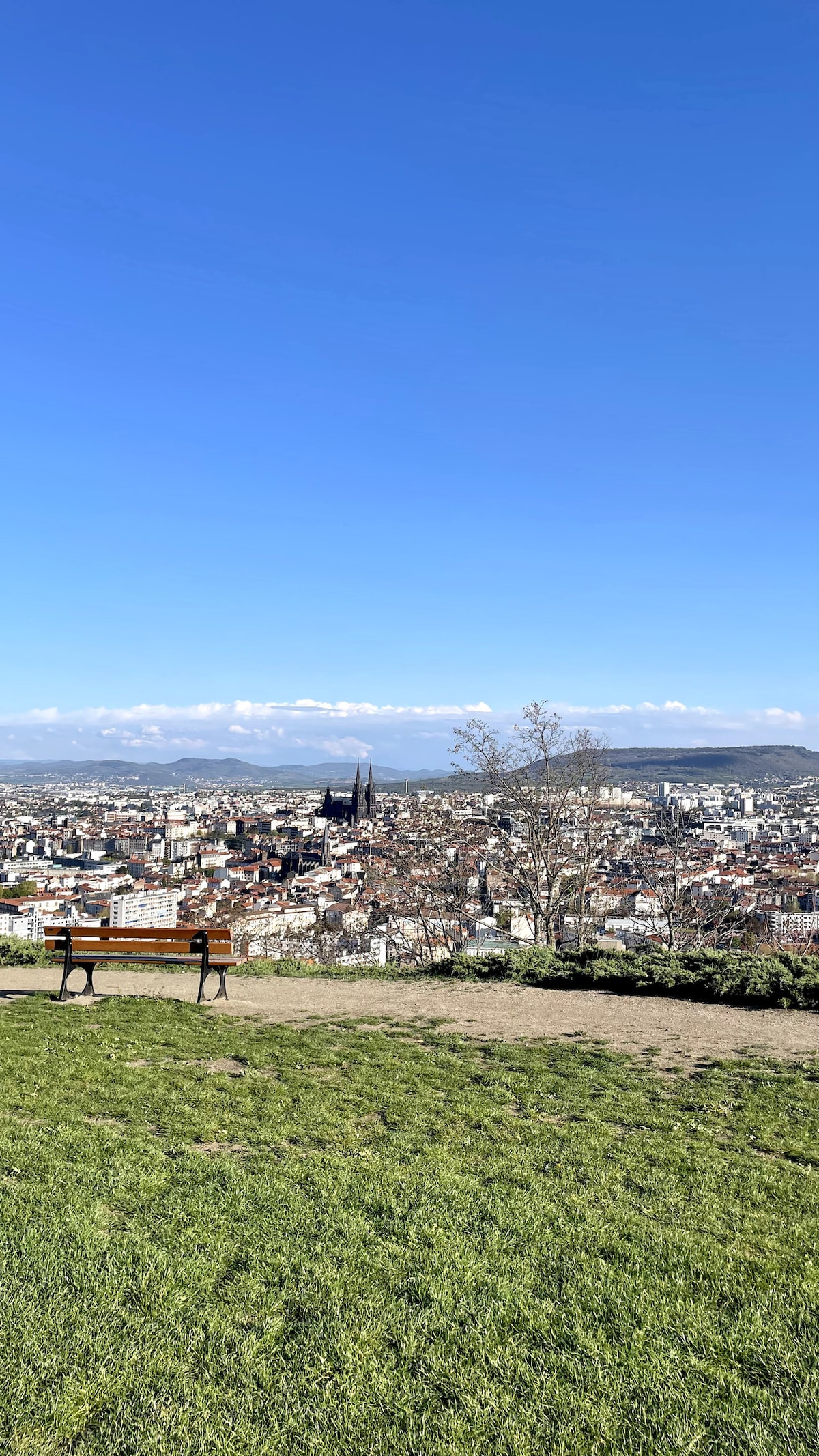 A clear view of Clermont-Ferrand is presented from a hillside, showcasing the city's architecture against a blue sky. A wooden bench is positioned on a grassy area, inviting viewers to enjoy the panoramic scenery of the city and surrounding landscape.