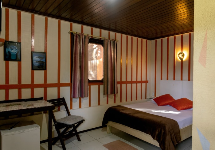 The room features rustic wooden paneling with orange and white stripes. A spacious bed with decorative pillows is complemented by a small dining table and a chair. Natural light enters through a window with curtains, and a small fridge is visible in the corner.