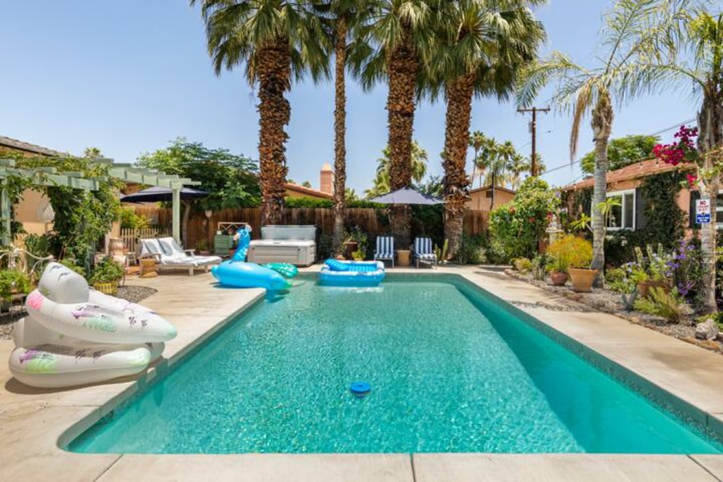 A serene outdoor space is highlighted by a large vintage pool surrounded by lush palm trees. Comfortable lounge chairs and umbrellas provide shaded seating areas. Colorful pool floats rest on the water, inviting relaxation in the sunny ambiance of Palm Springs.