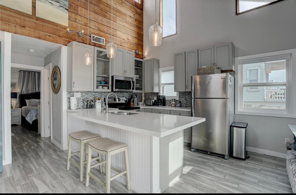 A modern kitchen is highlighted with sleek grey cabinetry and stainless steel appliances. A spacious kitchen island with two stools offers additional seating. Natural light enters through large windows, illuminating the open layout that connects to the living area.