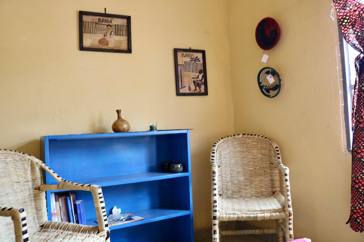 A pair of woven chairs is positioned around a vibrant blue shelf displaying books and decorative items. The walls feature framed artwork that reflects local culture, alongside handcrafted baskets hung in various colors. Natural light filters through the window with a patterned curtain.