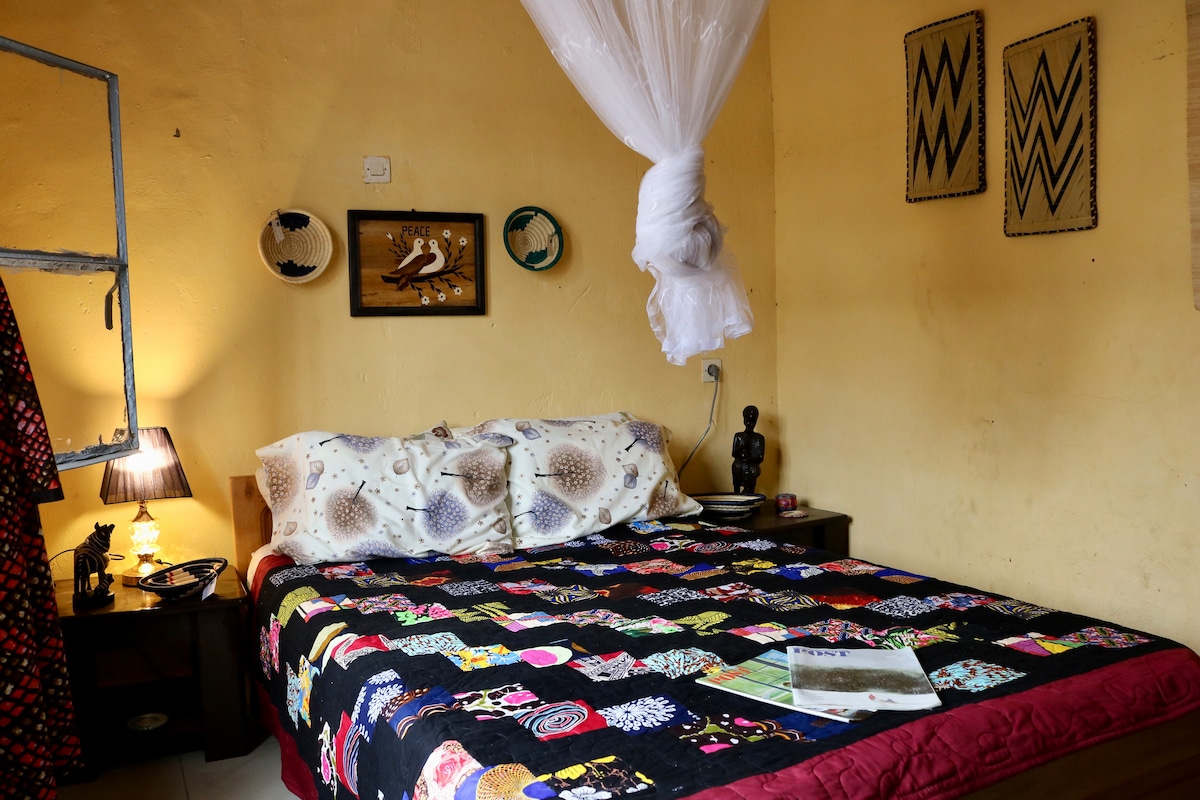 A cozy room features a spacious bed adorned with a colorful quilt. Handcrafted artwork decorates the walls, while a bedside lamp provides soft illumination. A small statue and decorative items are placed on the nightstand, creating a culturally rich atmosphere.