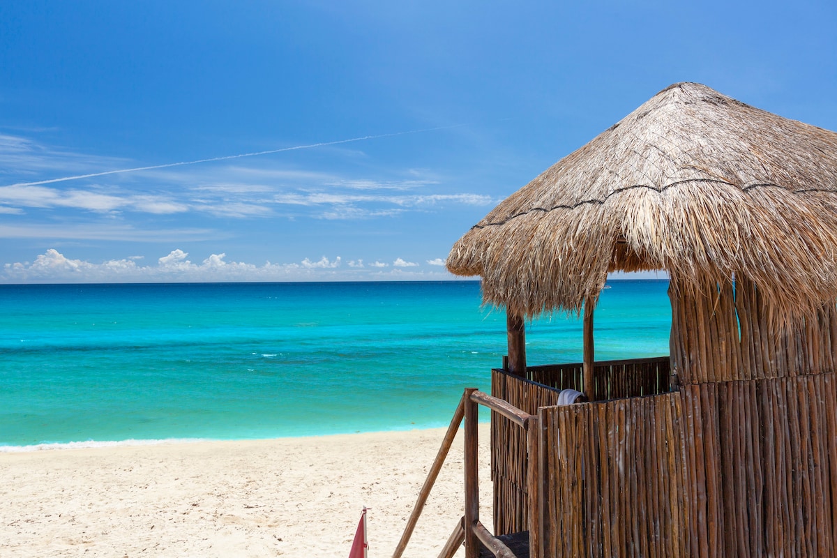 A thatched-roof structure is positioned on the beach, providing a shaded area that overlooks the turquoise waters of the ocean. The sandy beach extends in the foreground, with gentle waves lapping at the shore under a clear blue sky.