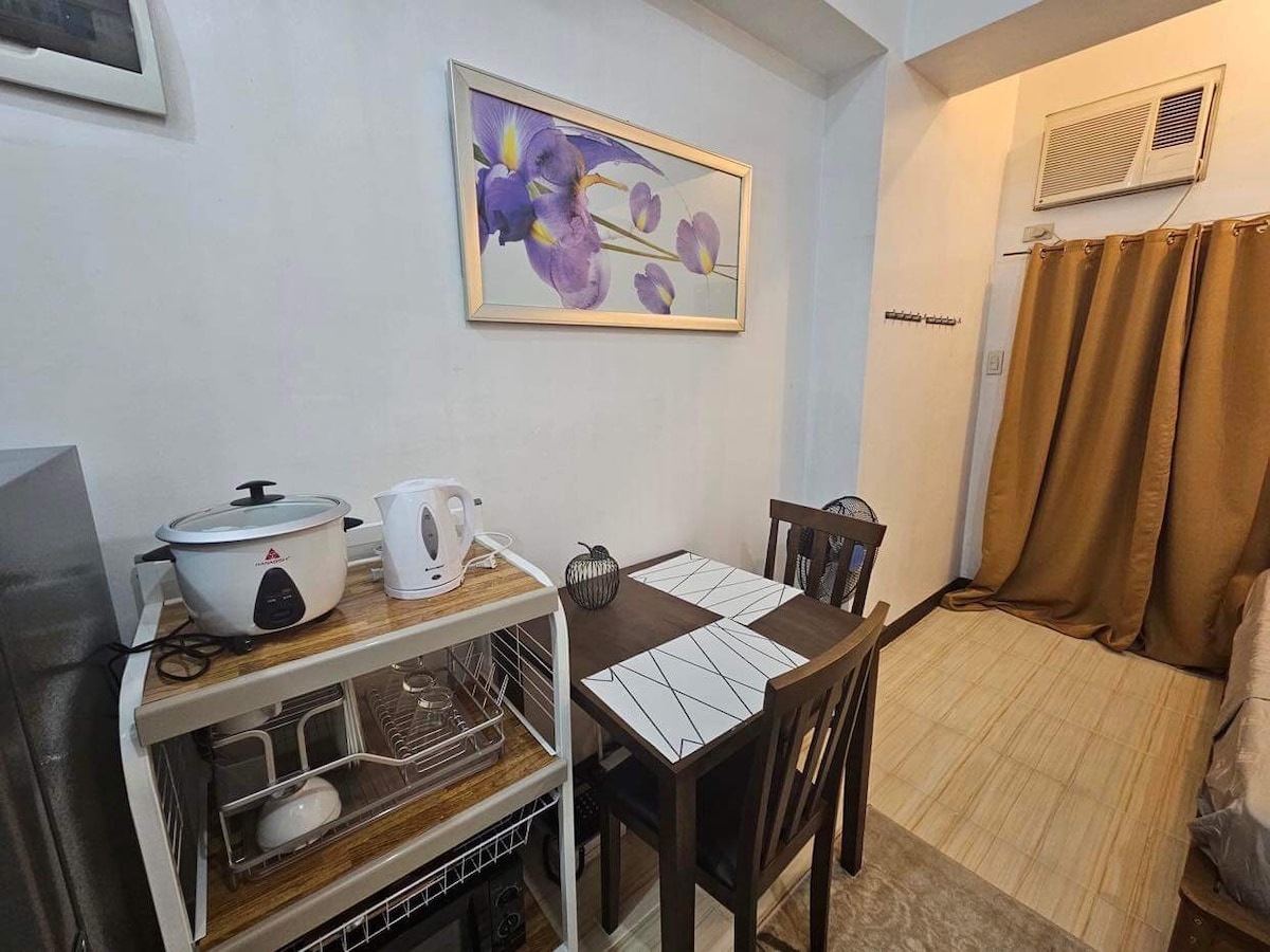 A small dining area is seen, featuring a table with two chairs and a floral artwork hanging above. A rice cooker and utensils are neatly arranged on a shelf next to a refrigerator, while a curtain separates the space from the sleeping area.
